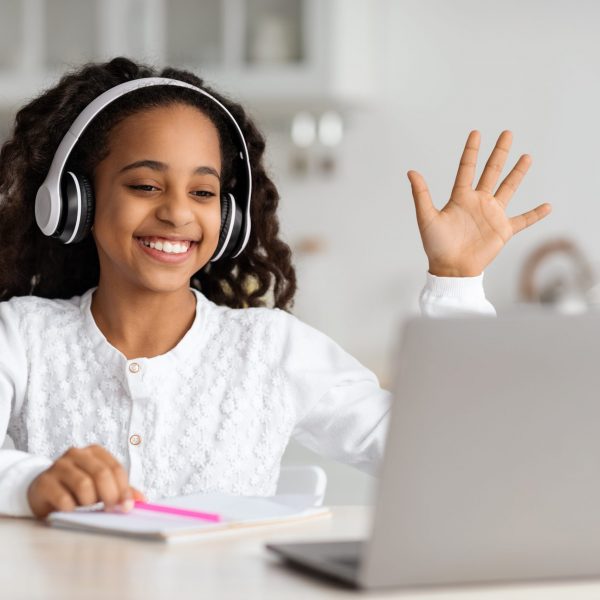 Positive african american kid cheerfully waving at laptop screen, using wireless headphones. Smiling black school girl having video chat with tutor, enjoying studying from home, kitchen interior